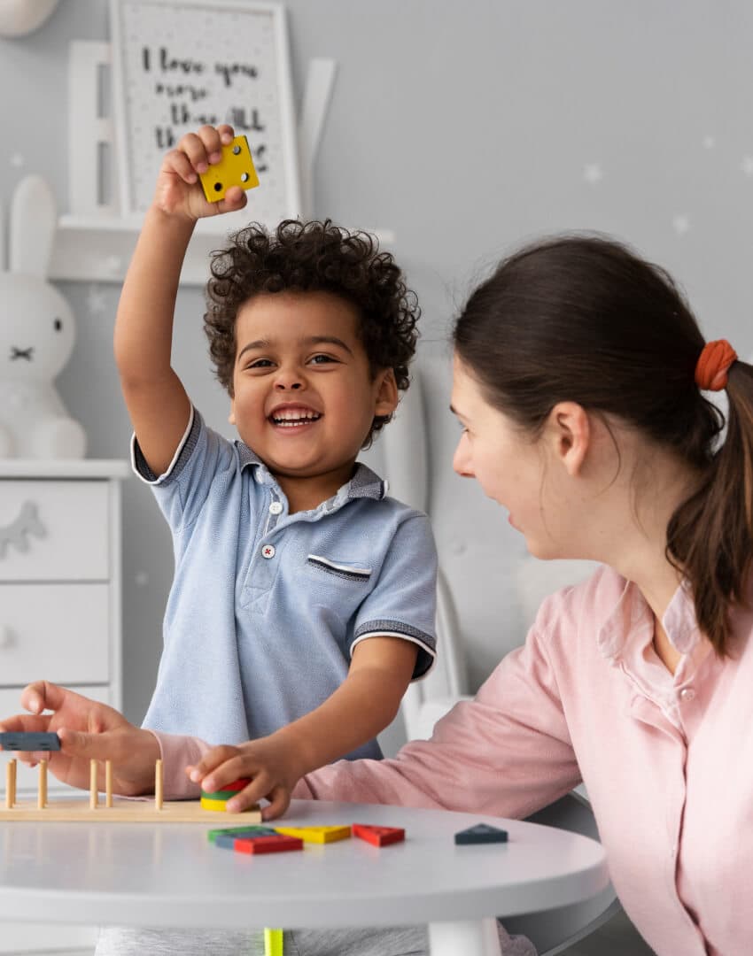 um menino de 3 anos de idade, sorrindo e mostrando uma peça de um brinquedo de encaixar, acompanhado de uma mulher adulta.